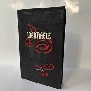 May include: A hardcover book titled "INSATIABLE" by Leigh Rivers. The cover is black with a red snake design over a skull and roses. The book is standing upright, showcasing the spine and front cover.