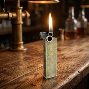 Pode incluir: Um isqueiro retangular de metal em tom de latão com um acabamento desgastado e envelhecido. O isqueiro apresenta uma abertura circular no topo e está aceso, exibindo uma chama vertical brilhante e constante que sai do mecanismo superior.