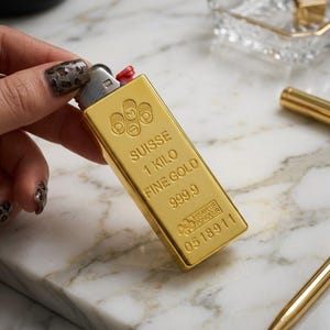 Puede incluir: Un encendedor rectangular de color dorado diseñado para parecerse a un lingote de oro suizo. La superficie presenta texto en relieve que dice SUISSE, 1 KILO, FINE GOLD, 999.9 y ESSAYEUR FONDEUR, junto al número de serie 0518911 y un logo.