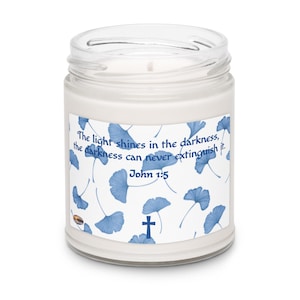 Puede incluir: Una vela en tarro de cristal blanco con tapa. La etiqueta presenta hojas de ginkgo azules y el texto "La luz brilla en las tinieblas, las tinieblas nunca podrán extinguirla. Juan 1:5" con una cruz.