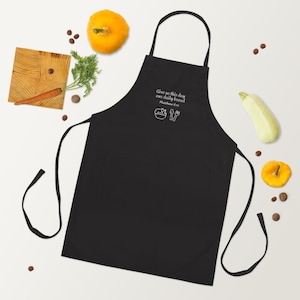 Op de afbeelding: Zwarte schort met een slab en twee zakken. De schort heeft een witte geborduurde tekst met de tekst "Geef ons heden ons dagelijks brood Mattheüs 6:11" met een kom, vork en mes. De schort ligt op een wit oppervlak met groenten.
