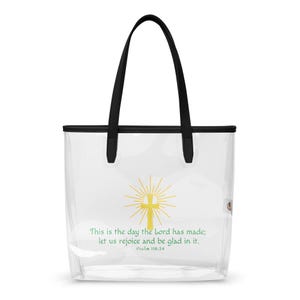 May include: Clear tote bag with black handles and trim. The bag features a gold cross design with radiating lines and the text "This is the day the Lord has made; let us rejoice and be glad in it. Psalm 118:24."