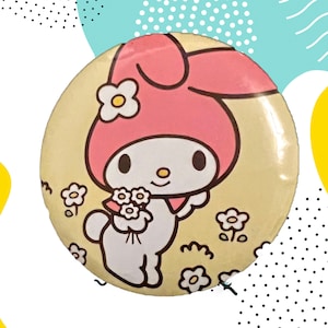 Pode incluir: Um botão redondo com um personagem de desenho animado com um capuz rosa e um corpo branco, segurando flores. O botão tem um fundo amarelo claro com flores brancas espalhadas e um contorno preto.