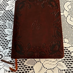 May include: A dark brown leather-bound book with a debossed floral border pattern along the edges. The center of the cover features the embossed text "The Holy Bible" in a script font. A thin brown ribbon bookmark extends from the bottom.
