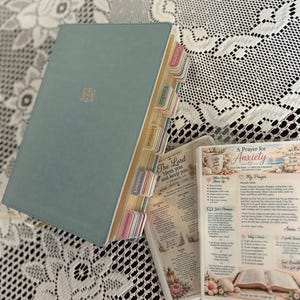 May include: A sage green Bible with gold embossed detail on the cover and multi-colored index tabs labeled with book names. Beside it lie two printed cards featuring religious text, including titles like A Prayer for Anxiety and The Lord.