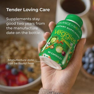 Op de afbeelding: Een groene fles van het voedingssupplement "Veggies", met een groene dop, in een hand. De fles toont afbeeldingen van groenten en de tekst "90 Capsules". De tekst "Tender Loving Care" en andere tekst zijn ook zichtbaar.