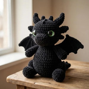 May include: A handmade black crochet dragon plush toy with large green eyes, wings, and horns. The dragon is sitting on a wooden surface near a window. The plush has a soft, textured appearance.
