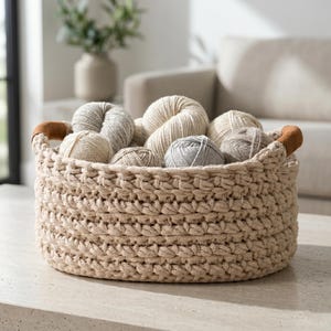 May include: A beige, crocheted basket filled with various yarn skeins. The basket has brown leather handles and sits on a light-colored surface. The yarn colors range from cream to gray, suggesting a craft or knitting theme.