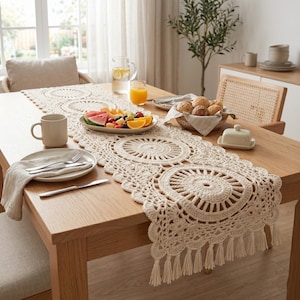May include: A beige crochet table runner with a circular design and fringe detail. The runner is placed on a wooden table set for a meal, with a plate of fruit, a mug, and silverware. A pitcher of water and a glass of juice are also on the table.
