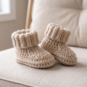 Può includere: Un paio di scarpine da neonato all'uncinetto beige fatte a mano. Gli stivaletti hanno un polsino a coste e un motivo a punto strutturato. Sono esposti su una superficie dai toni neutri, suggerendo un design accogliente e confortevole.