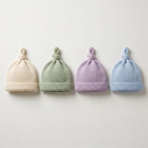 Könnte beinhalten: Vier gestrickte Baby-Mützen in Pastellfarben: Creme, Grün, Lavendel und Hellblau. Jede Mütze hat eine geknotete Oberseite und einen gerippten Saum. Die Mützen sind auf weißem Hintergrund in einer Reihe angeordnet.