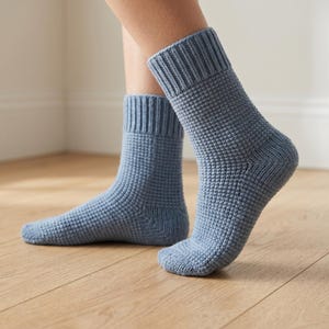 Può includere: Un paio di calzini lavorati a maglia blu chiaro. I calzini hanno un motivo strutturato e polsini a coste. I calzini sono mostrati sui piedi di una persona, con un piede leggermente sollevato. Sfondo di pavimento in legno chiaro.