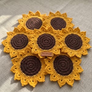 May include: A collection of six handmade crochet sunflower coasters. Each coaster features a brown center and yellow petals. A small, brown tag with the word "Handmade" is visible. The coasters are arranged on a neutral-colored surface.