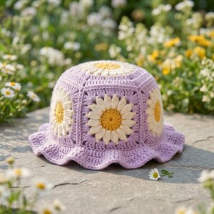 Pode incluir: Um chapéu de balde de crochê lilás com desenhos de flores de margarida. O chapéu apresenta um padrão quadrado com margaridas brancas e amarelas. A aba tem uma borda com babados. O chapéu está em uma superfície de pedra com flores ao fundo.