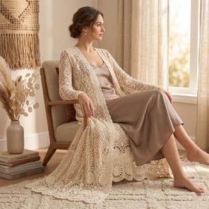May include: A cream-colored, open-front crochet cardigan with long sleeves and a floral pattern. The cardigan is draped over a person wearing a beige slip dress and sitting in a wooden chair. The room has neutral tones and natural light.