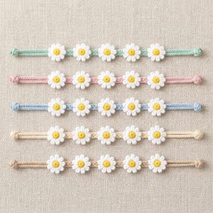 Könnte beinhalten: Fünf Gänseblümchen-Kettenarmbänder in Pastellfarben: Mintgrün, Rosa, Hellblau, Beige und Hellbraun. Jedes Armband hat gehäkelte weiße Gänseblümchen mit gelben Zentren. Die Armbänder sind horizontal angeordnet.