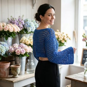 Könnte beinhalten: Ein blauer Häkeltop mit langen, ausgestellten Ärmeln. Das offene Design legt die Haut darunter frei. Das Top wird mit einer schwarzen Hose kombiniert. Der Hintergrund zeigt ein Blumengeschäft mit verschiedenen Rosen- und Hortensiensträußen.