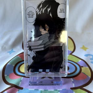 May include: A clear acrylic standee featuring a black and white manga illustration of a character with long dark hair and a scarf. The text reads: "LET'S TRY TO DO THINGS RIGHT THIS TIME, MIDORIYA" and "SO I'LL WATCH OVER YOU."