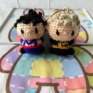 May include: Two small crochet dolls with round heads and black eyes. The left doll has black hair and a red, white, and blue outfit. The right doll has blonde hair and a black outfit with yellow accents. Both dolls feature small loops for hanging.
