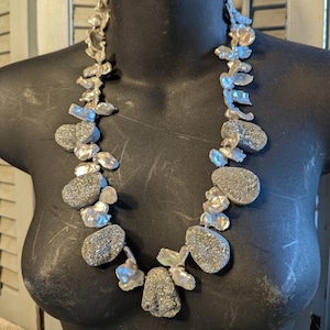 May include: A statement necklace featuring large, irregularly shaped silver-toned beads and iridescent, off-white baroque pearls. The necklace is displayed on a black mannequin, showcasing its unique design and texture. The necklace is a bold piece of jewelry.