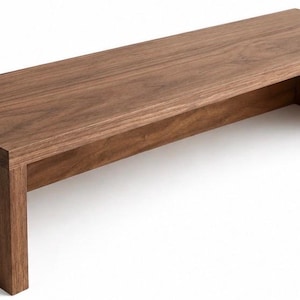 Peut inclure: Un banc rectangulaire en bois avec une finition au grain brun foncé lisse. La pièce présente un design en cascade continu où la surface supérieure se fond parfaitement dans deux supports latéraux verticaux, créant une structure ouverte.