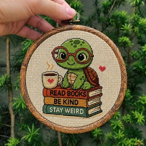May include: A cross-stitch embroidery hoop featuring a green turtle wearing glasses, sitting on a stack of books. The books read "READ BOOKS, BE KIND, STAY WEIRD." A cup of coffee with a heart is next to the turtle. The hoop is framed in wood.