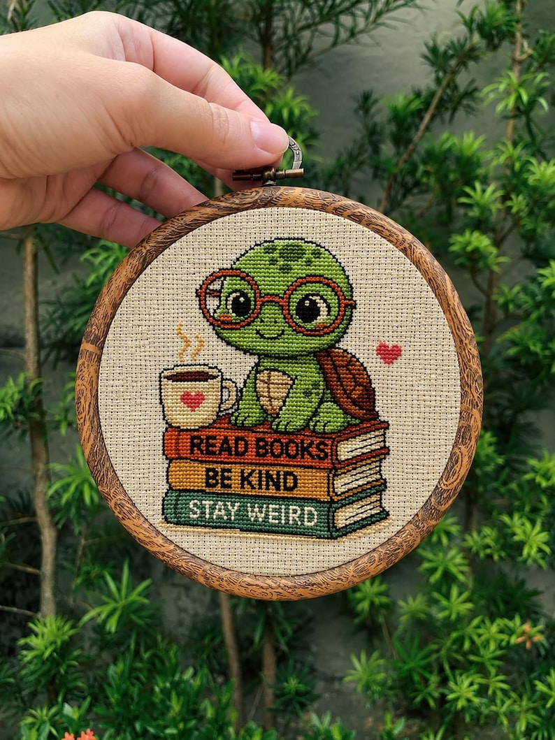 Op de afbeelding: Een borduurring met kruissteek met een groene schildpad met een bril, zittend op een stapel boeken. De boeken luiden "READ BOOKS, BE KIND, STAY WEIRD". Een kopje koffie met een hartje staat naast de schildpad. De ring is van hout.