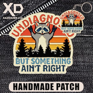 May include: An embroidered patch and a matching keychain featuring a raccoon with raised paws against a sunset and forest background. The text reads UNDIAGNOSED BUT SOMETHING AIN'T RIGHT. The design uses orange, yellow, blue, and black thread colours.