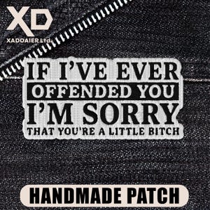 May include: A handmade patch with the text "IF I'VE EVER OFFENDED YOU I'M SORRY THAT YOU'RE A LITTLE BITCH" in black and white. The patch is rectangular with a white border and is set against a dark denim background.
