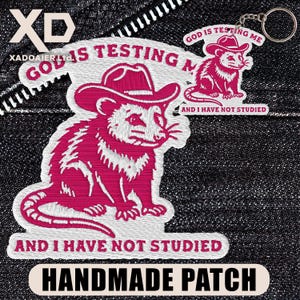 May include: Embroidered patch and keychain featuring a pink opossum wearing a cowboy hat. The patch and keychain have the text "GOD IS TESTING ME AND I HAVE NOT STUDIED" and "HANDMADE PATCH".