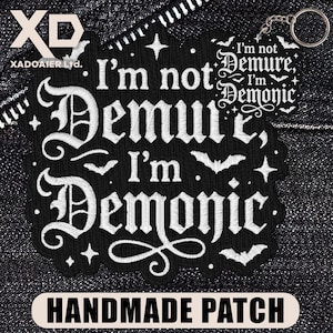 May include: Black embroidered patch with the text "I'm not Demure, I'm Demonic" in white gothic lettering. The patch features bats, stars, and the words "HANDMADE PATCH" at the bottom. A keychain is attached.