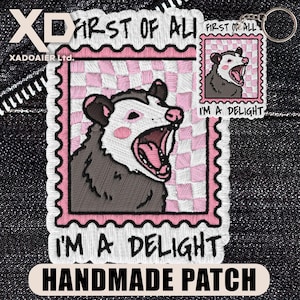 May include: Embroidered patch featuring a cartoon opossum with an open mouth, set against a pink and white checkered background. The patch reads "FIRST OF ALL" and "I'M A DELIGHT". A smaller keychain with the same design is also visible.