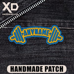 May include: An embroidered patch featuring a barbell design with blue and yellow thread. The center of the barbell includes the text ANYNAME in blue letters. The patch is displayed against a dark denim fabric background with a visible zipper.