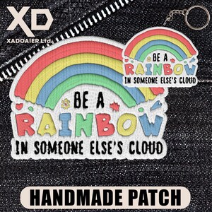 May include: Embroidered patch with a rainbow design and the text "BE A RAINBOW IN SOMEONE ELSE'S CLOUD." The patch features red, orange, yellow, green, and blue arches. The words "HANDMADE PATCH" are at the bottom.