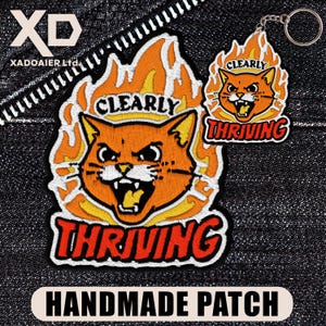 May include: Embroidered patch and keychain featuring an orange cat with the words "Clearly Thriving" in a flame design. The patch has the text "HANDMADE PATCH" at the bottom. The keychain has a silver ring.