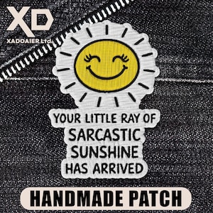 May include: Embroidered patch featuring a yellow smiling sun with eyelashes and radiating lines. The text "YOUR LITTLE RAY OF SARCASTIC SUNSHINE HAS ARRIVED" is below the sun. The patch is labeled "HANDMADE PATCH" on a black background.