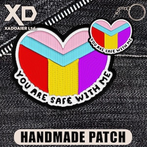 May include: A handmade heart-shaped patch with a white border, featuring red, yellow, purple, light blue, and pink sections. The text "YOU ARE SAFE WITH ME" is embroidered around the heart. A smaller keychain version is also visible.