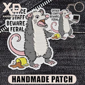 May include: Embroidered patch featuring a cartoon opossum with an open mouth, gray fur, and a pink tail. The patch includes the text "OFFICE STAFF BEWARE FERAL" and "HANDMADE PATCH". A keychain with a similar design is also visible.