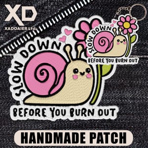 May include: A handmade patch with a cartoon snail design, pink shell, and the text "SLOW DOWN BEFORE YOU BURN OUT." The patch includes a flower and hearts, with a keychain attached. The patch is on a denim background.