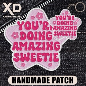 May include: A pink star-shaped handmade patch with the text "YOU'RE DOING AMAZING SWEETIE" in bright pink. The patch has a white border and small flower details. A smaller star-shaped keychain with the same text is attached.