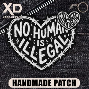 May include: A black and white embroidered patch in the shape of a heart with the words "NO HUMAN IS ILLEGAL". The patch is surrounded by barbed wire and the words "HANDMADE PATCH" are at the bottom.