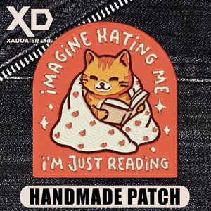 May include: Embroidered patch with a cartoon cat reading a book, wrapped in a blanket with heart details. The patch is orange with the text "IMAGINE HATING ME, I'M JUST READING" and "HANDMADE PATCH".