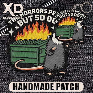 May include: Embroidered patch featuring a cartoon opossum next to a green dumpster engulfed in flames. The text reads "THE HORRORS PERSIST BUT SO DO I." A smaller keychain version is also visible. The patch is labeled "HANDMADE PATCH."