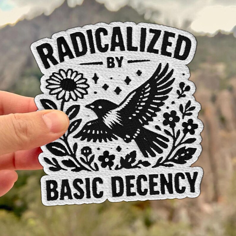 Peut inclure: &Eacute;cusson brod&eacute; avec le texte "RADICALIZED BY BASIC DECENCY" en noir sur fond blanc. L'&eacute;cusson repr&eacute;sente un oiseau noir en vol, entour&eacute; de fleurs et d'&eacute;toiles.