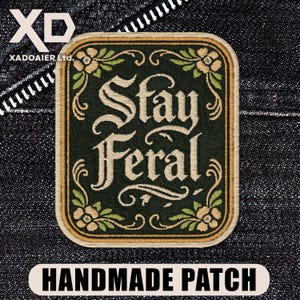 May include: A rectangular embroidered patch with rounded corners featuring a dark green background and the text Stay Feral in a cream-coloured gothic font. The border is tan with decorative floral accents in cream and light green at the corners.