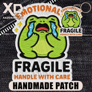 May include: Embroidered patch featuring a crying green frog with the text "EMOTIONAL" and "FRAGILE HANDLE WITH CARE". A smaller keychain version of the frog is also visible. The patch is handmade.