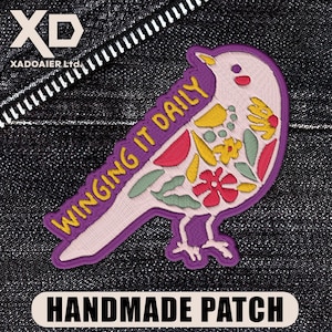 May include: A handmade patch in the shape of a bird, featuring the text "WINGING IT DAILY." The bird design includes colorful floral and leaf details in red, yellow, and green, outlined in purple. The patch is on a dark denim background.