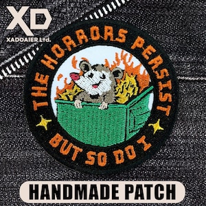 May include: Embroidered patch with a cartoon opossum in a green dumpster engulfed in flames. The text reads "THE HORRORS PERSIST BUT SO DO I." The patch is round with a black border and yellow stars. The bottom text says "HANDMADE PATCH."