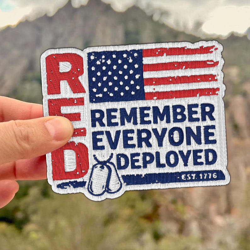 Pode incluir: Patch bordado com o texto "RED" &agrave; esquerda e um design da bandeira americana no canto superior direito. O texto "REMEMBER EVERYONE DEPLOYED" est&aacute; abaixo, com etiquetas de identifica&ccedil;&atilde;o e "EST. 1776" na parte inferior. O patch &eacute; segurado por uma m&atilde;o.