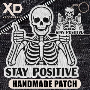 May include: Embroidered patch and keychain featuring a skeleton giving a thumbs-up. The patch reads "STAY POSITIVE HANDMADE PATCH". The keychain has a smaller skeleton design with the same message. Both items are white on a black background.